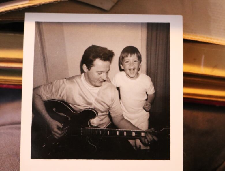 A black and white photo of a man playing electric guitar with a young boy watching and smiling. 