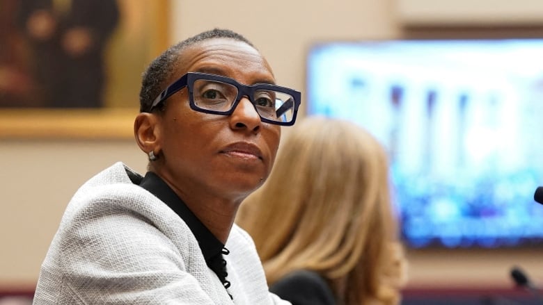 A woman with short cropped hair and thick black glasses watches a video on a big screen with her hands folded on the desk in front of her.