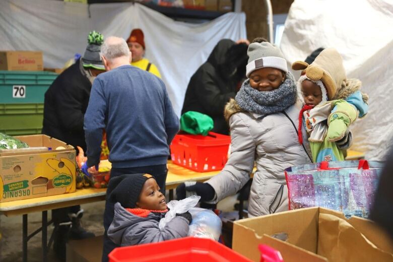 A woman carrying a baby hands another child a bag of food. They are all bundled up in winter clothes. 