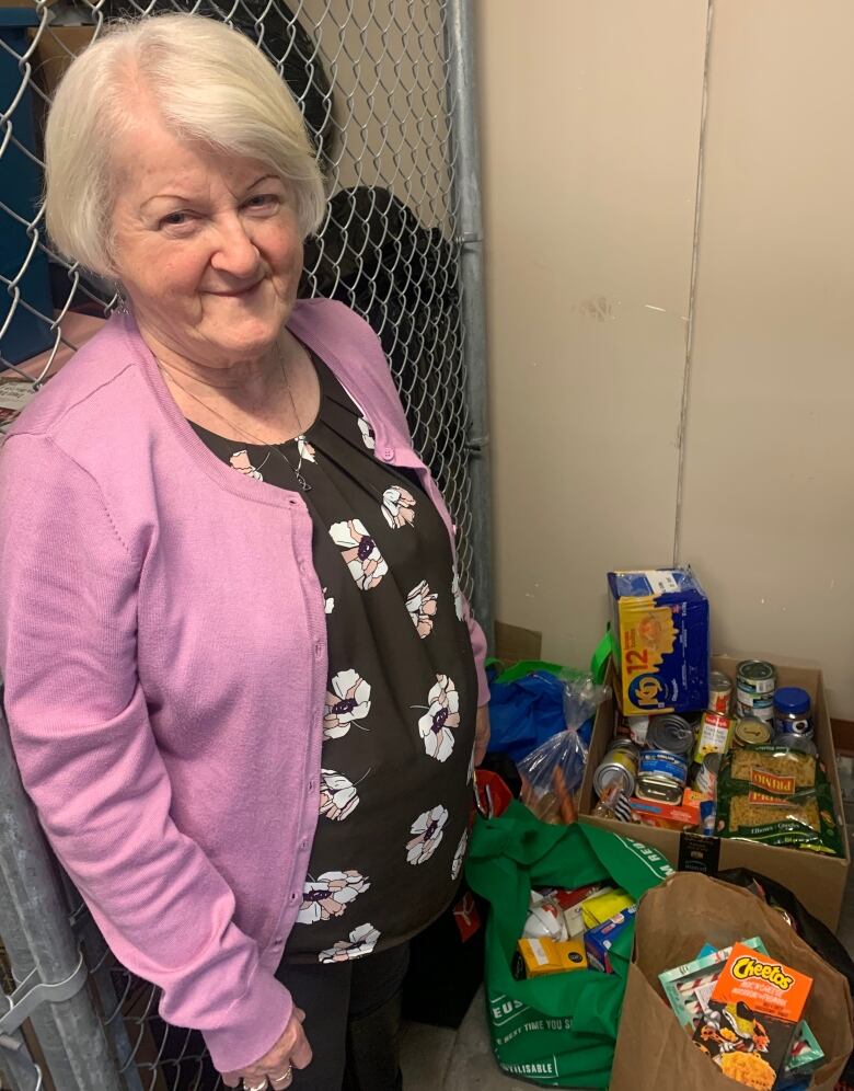 A senior lady, dressed in a pink cardigan, stands next to a few bags and boxes that are full of donated perishable and non-perishable food.