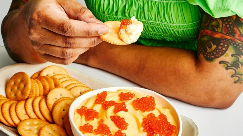 Closeup on a bowl of light yellow dip with orange trout roe on top. The bowl is sitting on a platter with Ritz crackers next to it. Behind it, a person wearing a lime green dress scoops into the dip with a cracker.