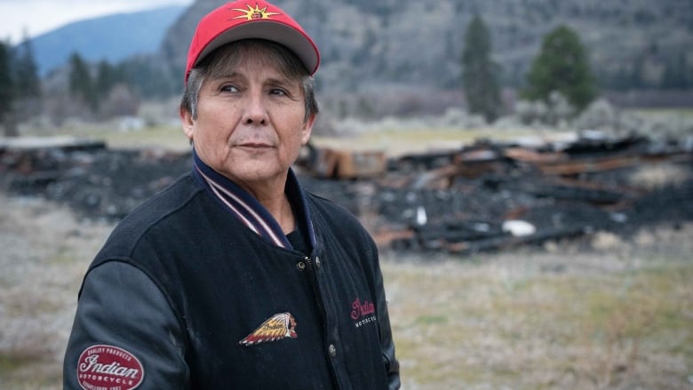 Indigenous man, red hat and black jacket stands in front of pile of charred debris.