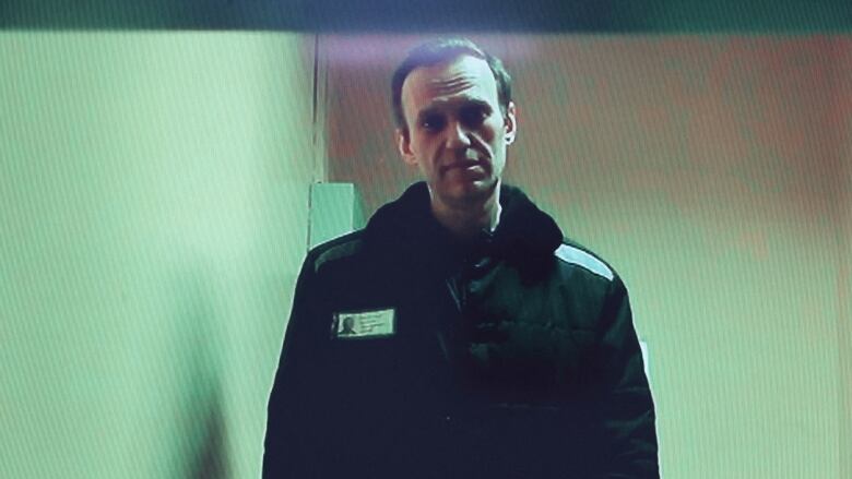 A man in a winter jacket stands in small, plain room, visible through a slightly blurry and green-hued screen.