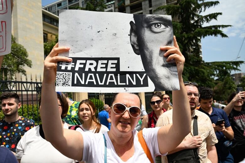 A woman in sunglasses stands in a crowd holding a sign with a man's face, a QR code and the words #FREE NAVALNY