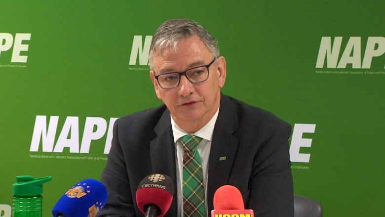 A man wearing a black suit sits in front of a green backdrop with the word 