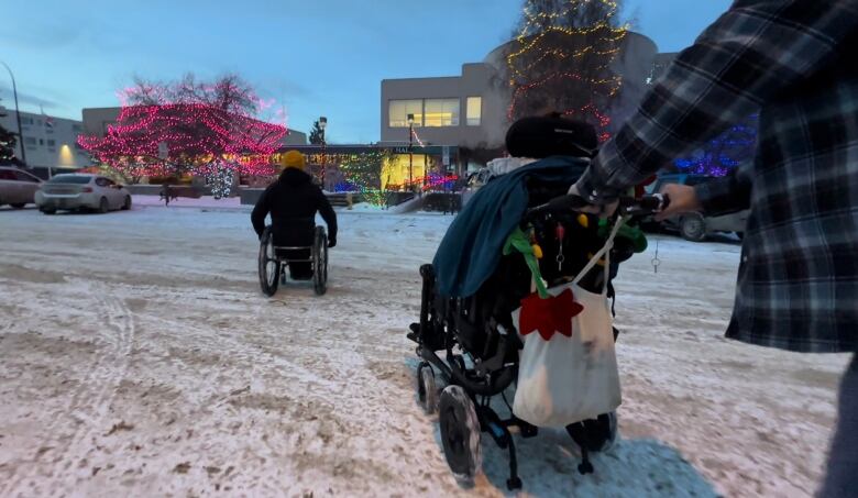 Two people in wheelchairs make their way across a snowy road.