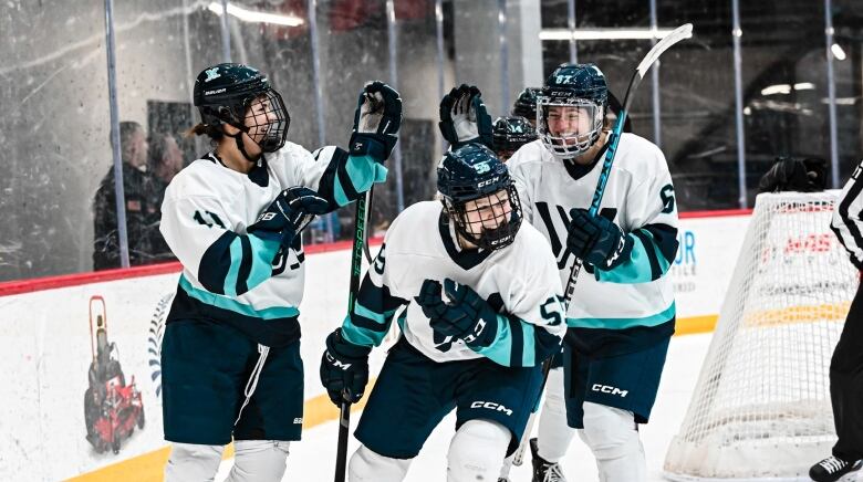 Three female hockey players in white and teal jerseys celebrate on the ice.