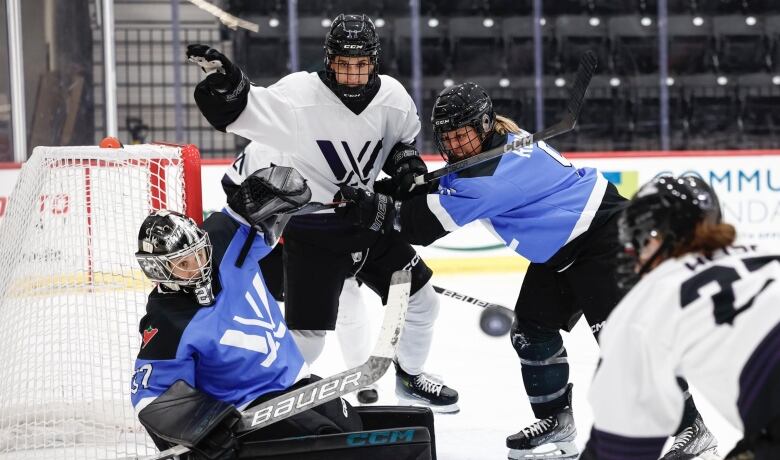 A female hockey player in a white jersey battles for the puck in front of the opposing team's net net with a teammate and a player from the opposing team.