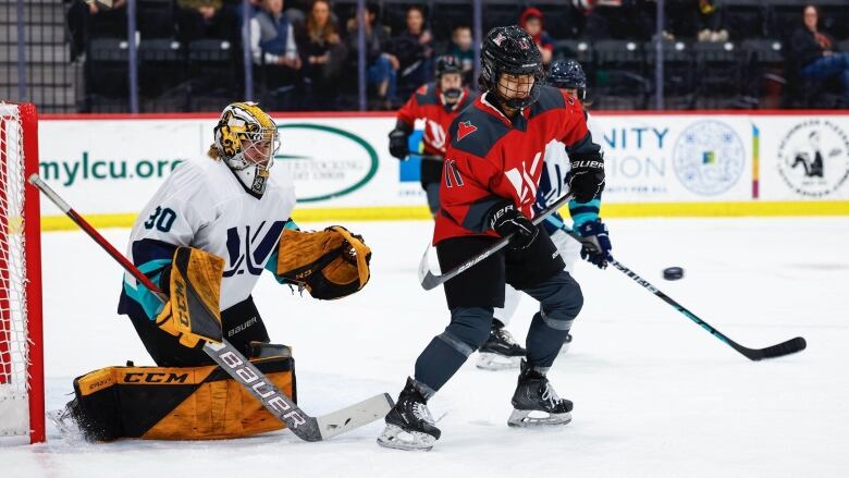 A female hockey player in a red jersey looks at the puck in front of the opposing team's net.