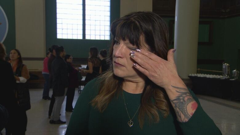 A woman is wiping away a tear from her left eye. She is standing in a room with people in the background.