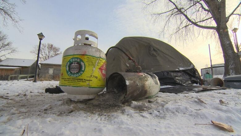 A propane tank is sitting in the snow. A metal cylinder is tipped over with ash spilling out of it. A tent, trees and a wooden house are in the back.