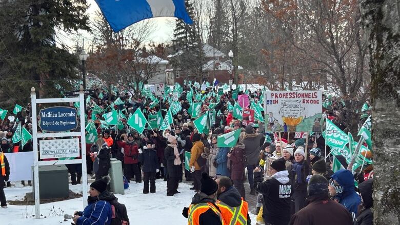 A crowd rallies outside a politician's office in winter. Many are waving teal flags.