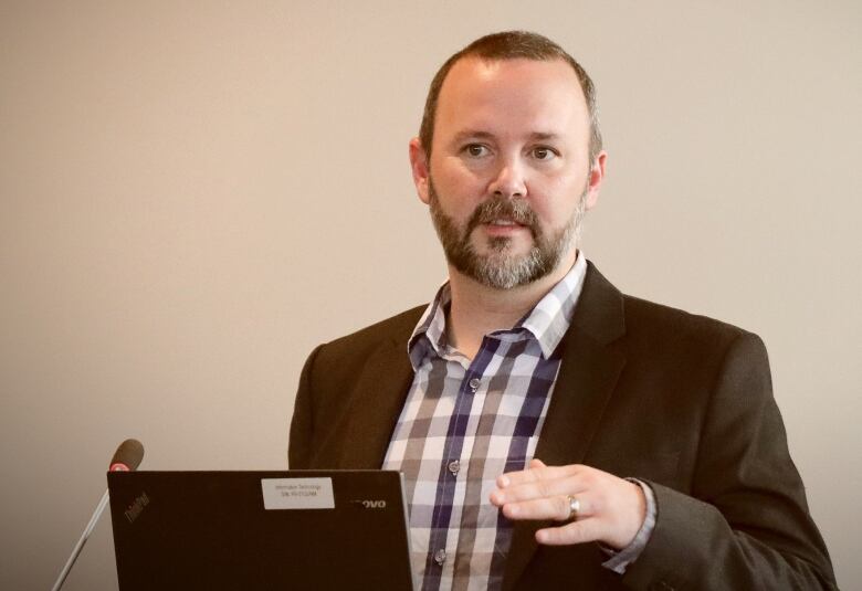 A man with beard and moustache, short hair and a brown suit jacket and a white-and-checked shirt speaks in front of a microphone and computer screen.