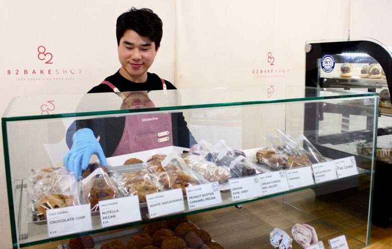 A young man wearing a pink apron organizes cookies in a glass display case at a bakery counter.