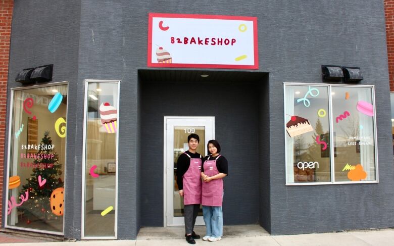 A young Korean man and woman wearing pink aprons stand in front of a store with a sign that says '82Bakeshop' over the door. The windows are dcecorated with decals of desserts.