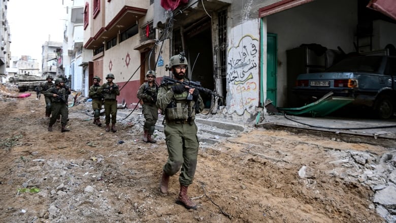 Six Israeli soldiers in green uniforms carry guns as they walk through a ruined street in Gaza.