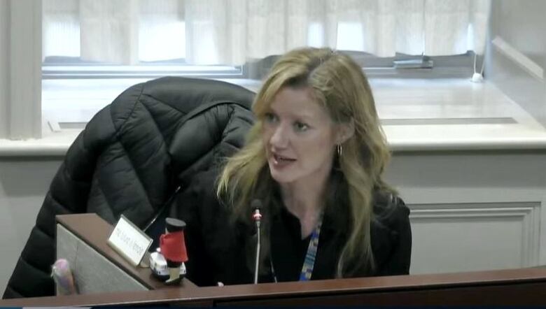 A white woman with long blonde hair and a black blouse sits at a desk in an office chair with a microphone in front of her