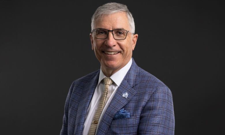 A man with glasses, salt-and-pepper hair and wearing a blue suit with brown tie smiles for the camera.