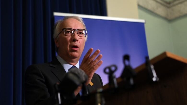 A middle-aged man stands at a podium wearing a black suit and black-framed glasses.