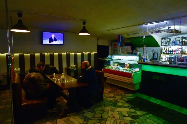 Two people sit around a small table in a dimly lit room with a bar. One looks up at a TV on the wall that shows Vladimir Putin.