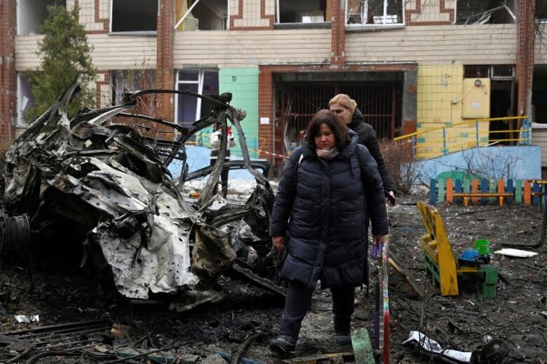 People walk beside a car that has been destroyed and a building that has been damaged.