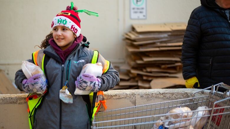 A girl stands wearing a christmas hat holding frozen chickens.