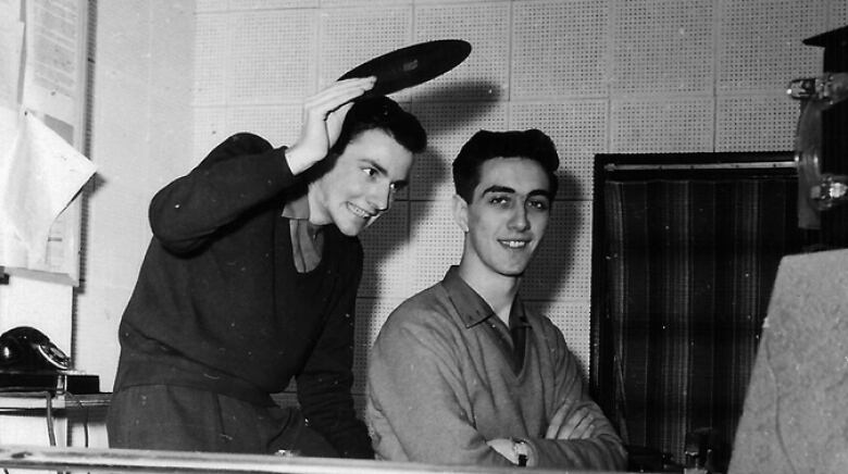 a black and white photo shows a young man holding a record over another man's head in a radio studio in the 1950s.