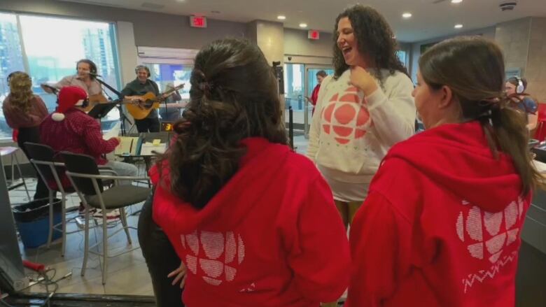 A woman in a white CBC logo sweater talks to two women in red CBC logo sweaters. 