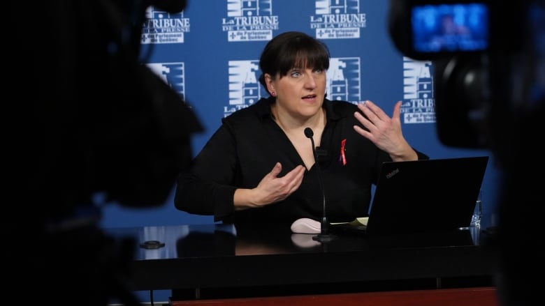 A white woman with bangs speaks to reporters in front of a white and blue background. With a laptop in front of her. 