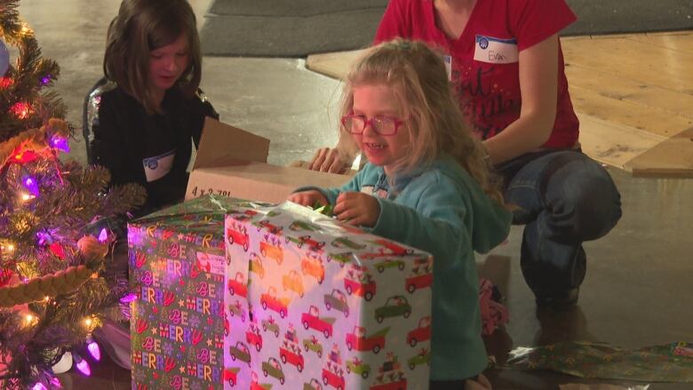 A little girl opens presents next to a Christmas tree. 