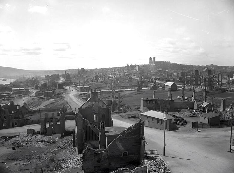 Rubble is seen on both sides of a street, with crumbling brick work in the foreground. Some smoke is rising from parts of a destroyed city. 