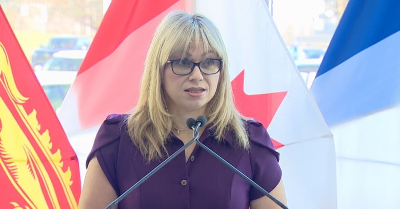 Woman with glasses and shoulder-length blond hair stands in front of microphones with flags in the background. 