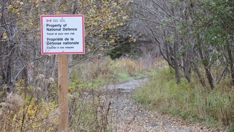 A white sign with black and red writing says Property of National Defence in front of a road that leads off into the woods.