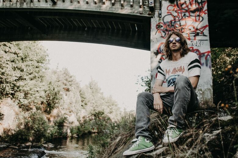 A man with long brown hair and mirrored sunglasses, wearing a white Def Leppard T-shirt and green sneakers, sits on the banks of a stream running under a bridge.