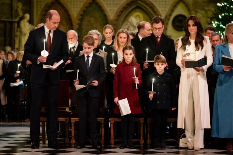 Three children stand between two adults as they hold candles and programs during a carol service in a large church.