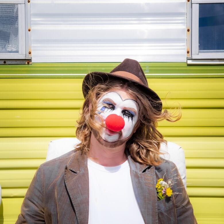 A man in a brown fedora has white clown makeup on his face, including a bright red clown nose.