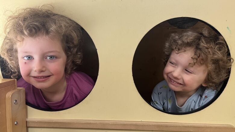 Two curly haired children stick their faces through holes in a play structure.