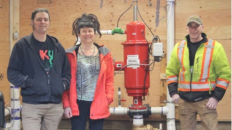 On the left is a man in a blue jacket with his hands in his pockets and a woman in a pink jacket with her hair up in a bun. On the right is a man wearing a high visibility jacket and a series of pipes between everyone. 