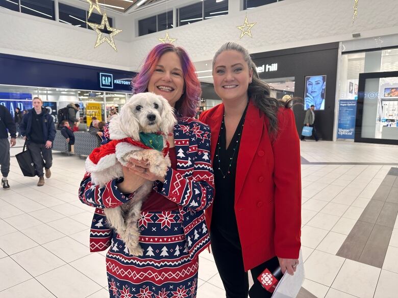 Woman in tree print suit holding a little white dog next to woman in red suit.