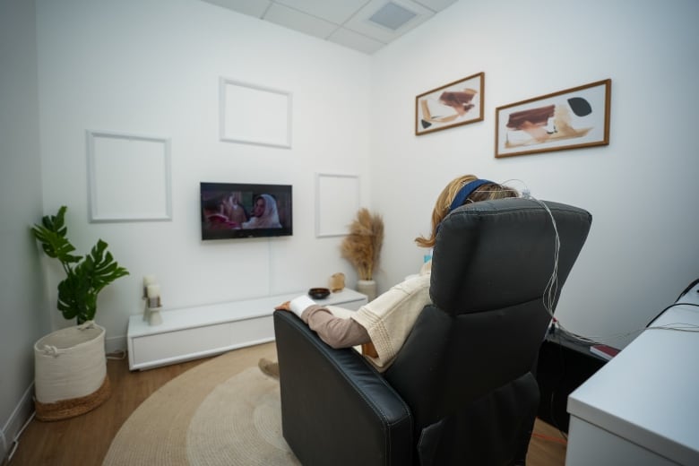 A woman sits in an armchair watching a film with earphones on, as her head is connected to a set of cables behind her.