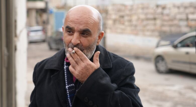 An elderly man, with a greyish beard and a bald head, wearing a dark overcoat, smokes a cigarette.