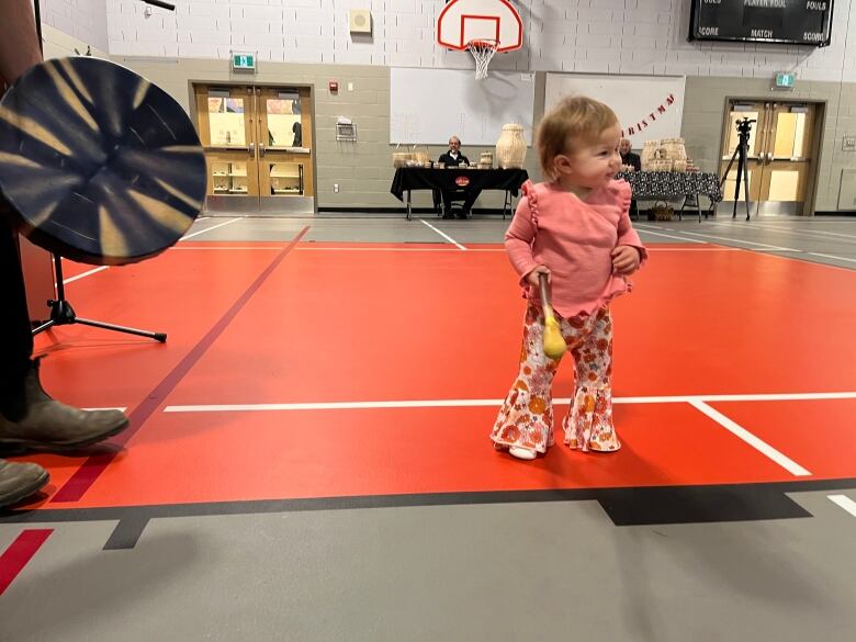 A toddler in a pink top and bell bottom pants holds a drum stick, while the drum is visible to her left.