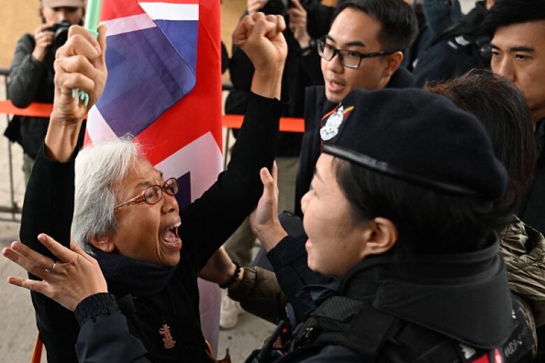 A woman with grey hair waves a flag and raises her fist in the air as a police officer, facing her, holds her arms. 