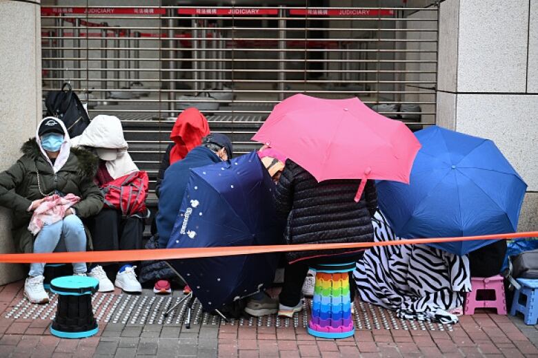 People huddle on stools and under umbrellas in a lineup outside a building.