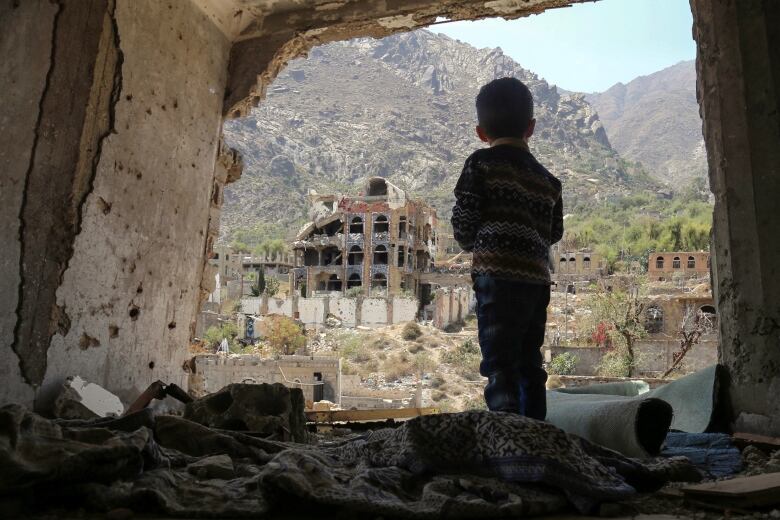 A boy with his back to the camera looks out on mountainous landscape with heavily damaged buildings in the foreground.