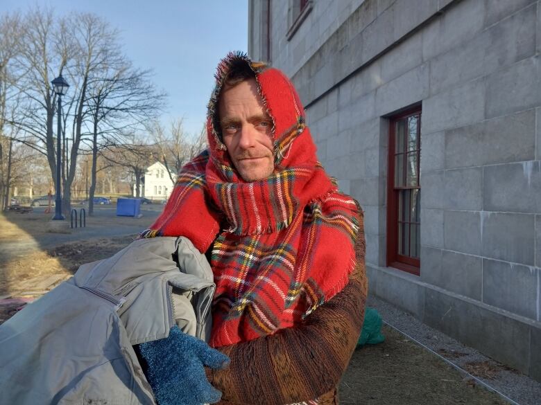 Man outdoors, bundled up in blankets. He has a red tartan scarf wrapped around his head and shoulders.