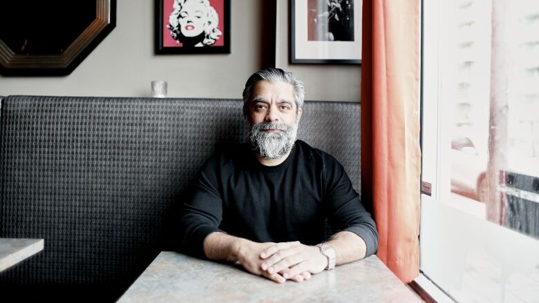 A man with a beard, wearing a longsleeve black t-shirt, sits by the window in a restaurant.