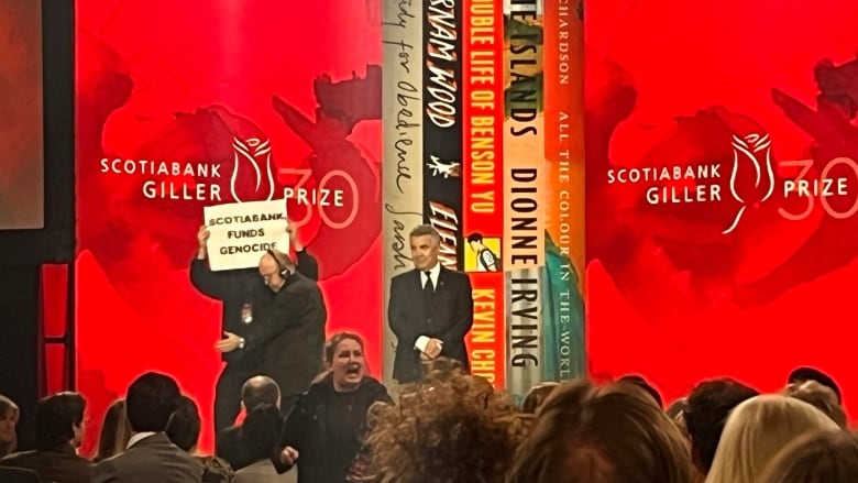 A person holding a sign that reads: Scotiabank funds genocide is escorted from the stage at an awards show, with another protester in the seated crowd shouting.