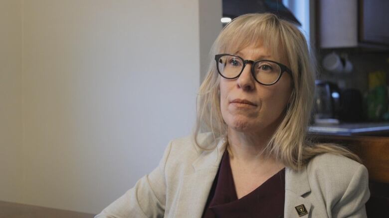 A woman with glasses, bangs and long hair is wearing a blazer with a v-neck top underneath.  