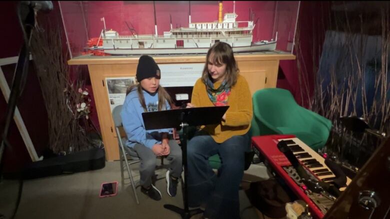 An 11-year-old girl with a toque and long dark hair sits beside a woman who's pointing to something on a music lectern.